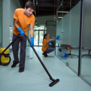 Young man vacuuming the floor while his collegue cleaning the glass surface in the building