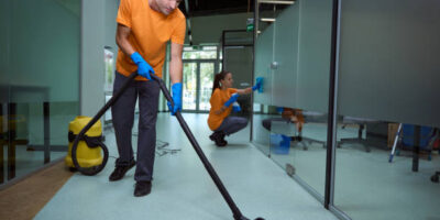 Young man vacuuming the floor while his collegue cleaning the glass surface in the building
