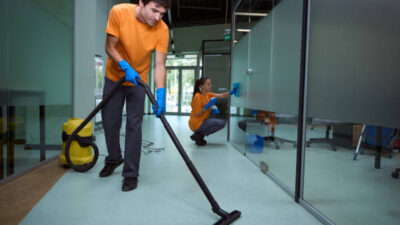 Young man vacuuming the floor while his collegue cleaning the glass surface in the building