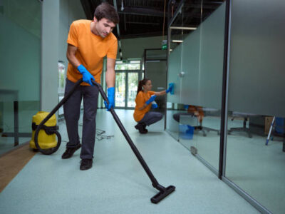 Young man vacuuming the floor while his collegue cleaning the glass surface in the building