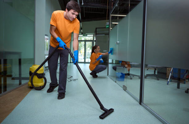 Young man vacuuming the floor while his collegue cleaning the glass surface in the building